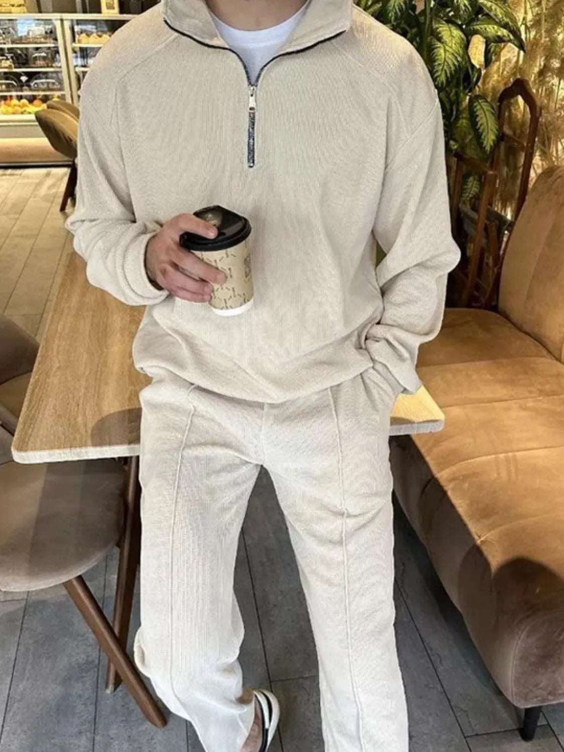 Person wearing a beige tracksuit holding a coffee cup in a casual setting.
