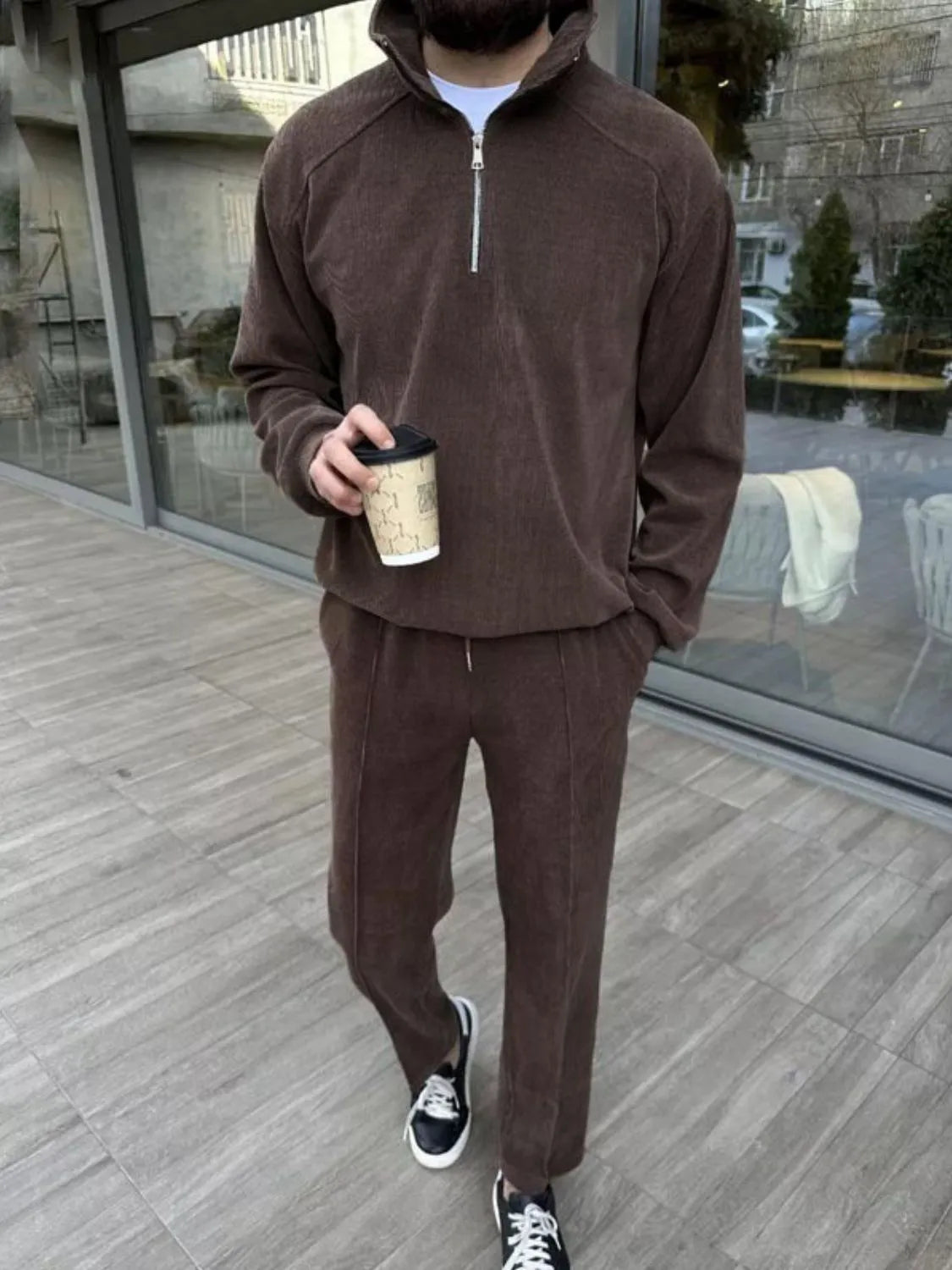 Person wearing a brown tracksuit holding a coffee cup on a wooden deck.