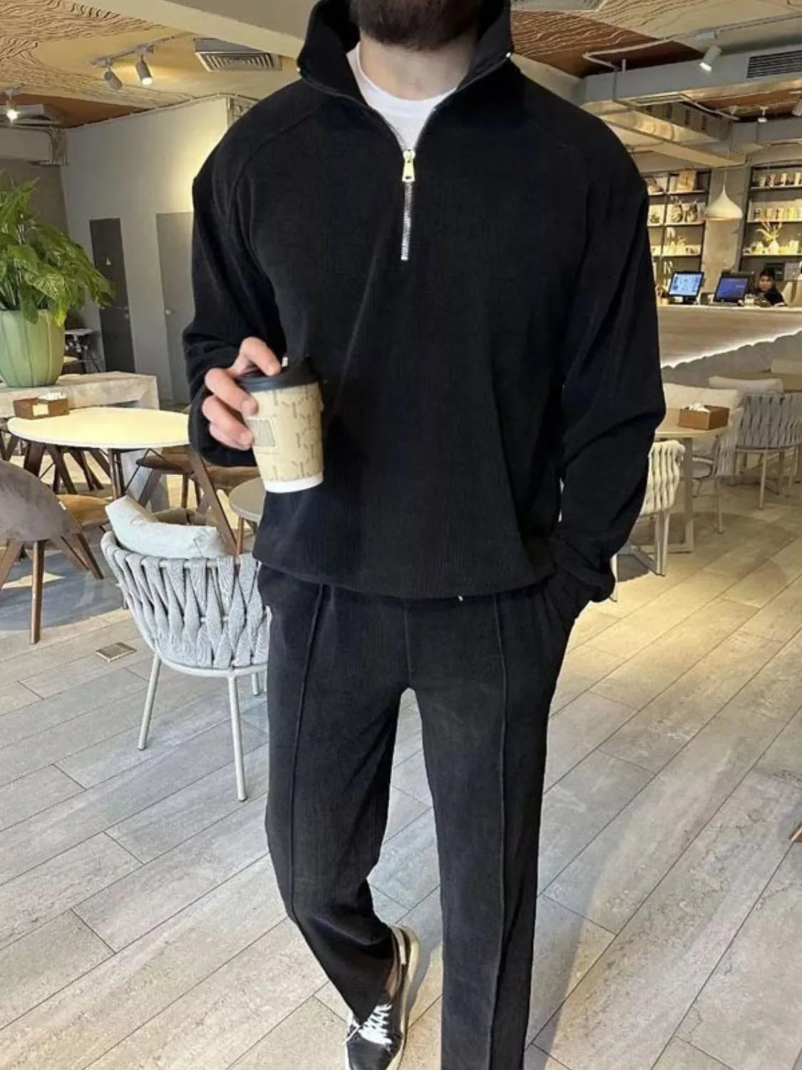 Person wearing a black tracksuit holding a coffee cup in a cafe.