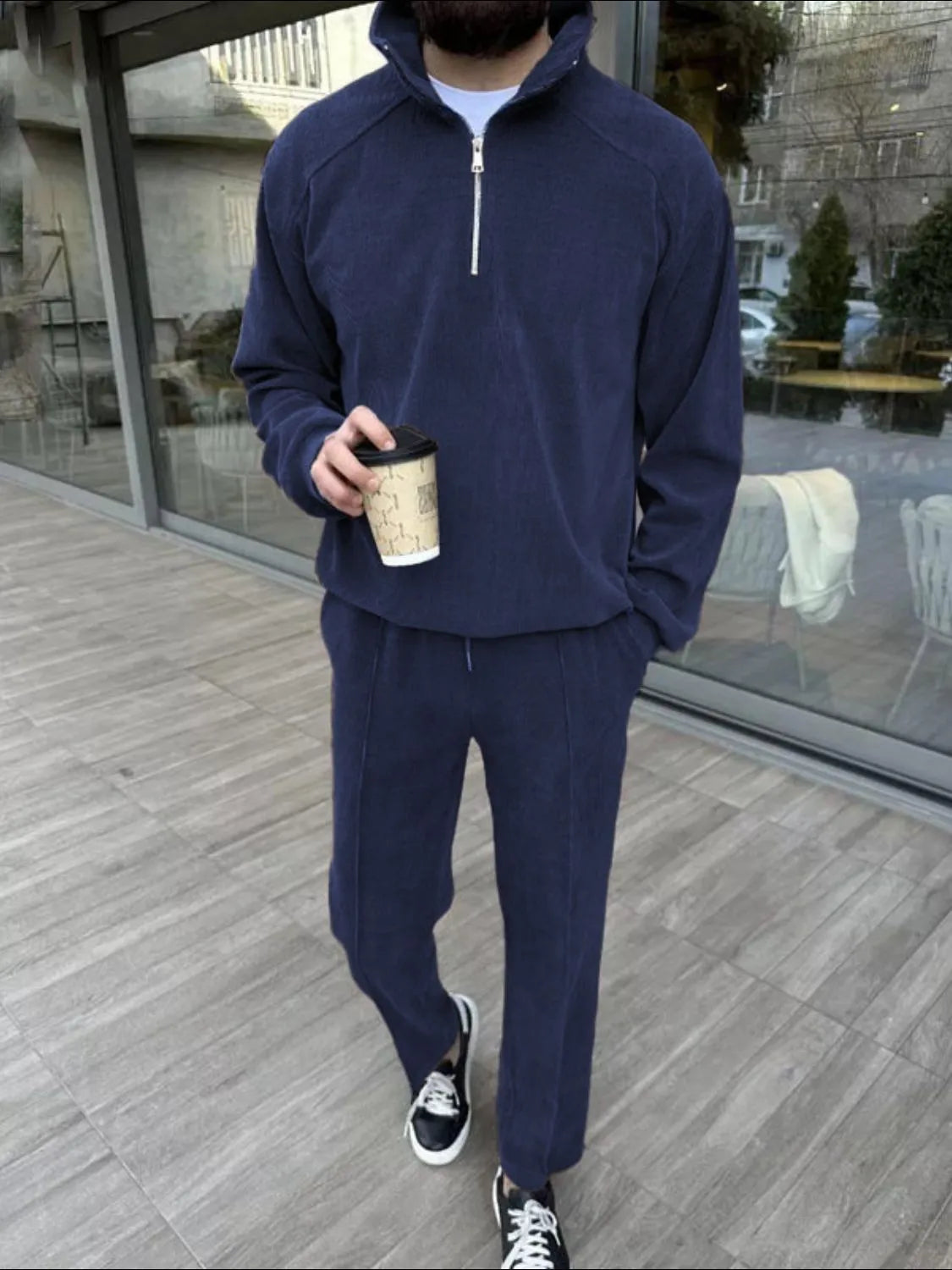 Person wearing a navy tracksuit holding a coffee cup on a wooden deck.
