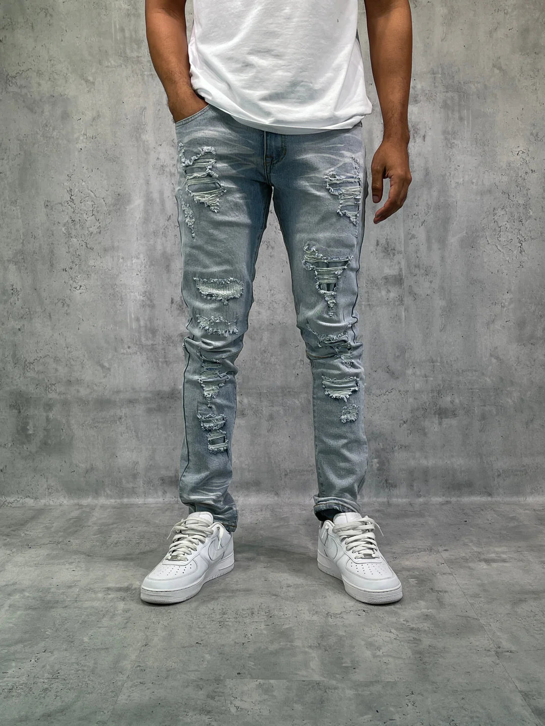 Man wearing light blue distressed jeans and white sneakers on a gray concrete background