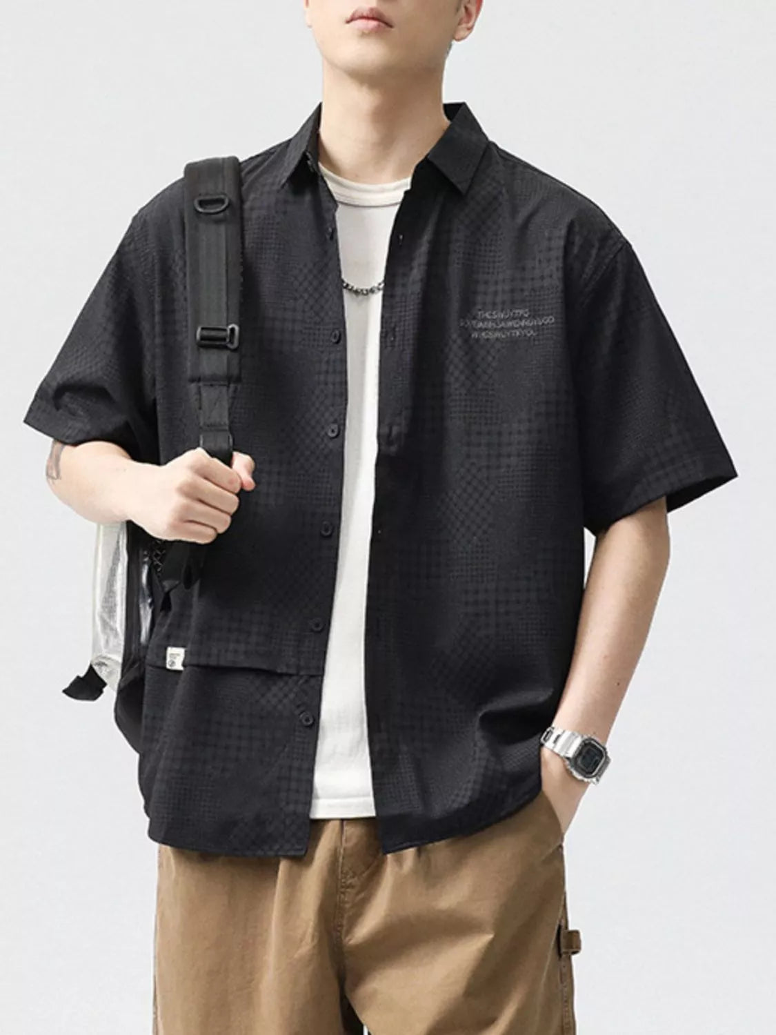 Person wearing a black textured shirt with a white t-shirt underneath, holding a backpack on a light gray background.